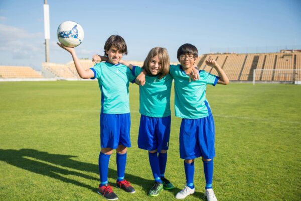 Three young soccer players in turquoise uniforms pose and smile on a sunny field in Portugal.